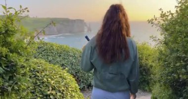 Güneş gözlüklü bir kadın ve bir uçurumun kenarında elbisesi Etretat sularındaki ünlü kayaların arkasında. Gün batımında uzun kıvırcık saçlı çekici bir kadın..