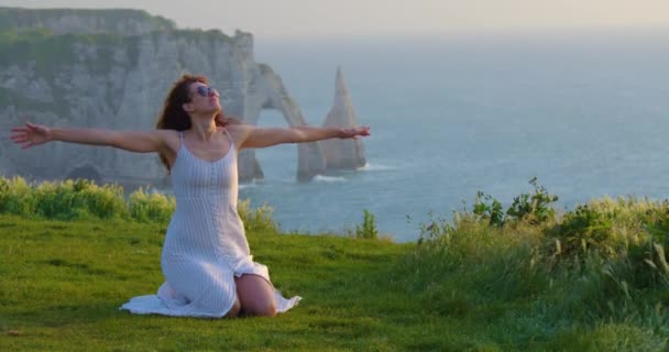 ビーチの背景に美しいドレスを着た女の子 夕日が女の子の顔に輝いています 大西洋に囲まれたフランスの海岸 緑の草原とフランスの高い崖 — ストック動画