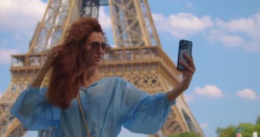 Parisli bir kadın akıllı telefondan fotoğraf çekiyor. Paris 'te Eyfel kulesi parkında ve caddelerde şık kıyafetler giyen genç bir kadının sinematik görüntüleri. Avrupa turizmi kavramı.