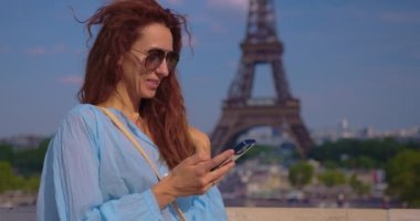 Akıllı telefon kullanan Parisli kadınlar. Paris 'te Eyfel kulesi parkında ve caddelerde şık kıyafetler giyen genç bir kadının sinematik görüntüleri. Avrupa turizmi kavramı.