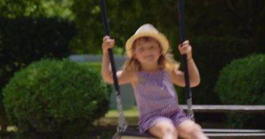 Üç yaşında, şapkalı ve elbiseli bir kız yazın parkta salıncakta sallanıyor..