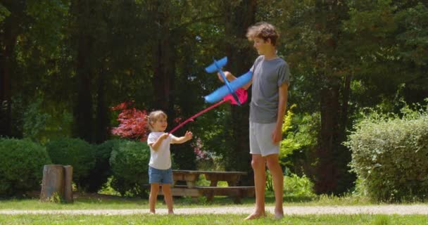 赤い網の赤ん坊は夏の緑の公園のティーネージャーと遊ぶ 青いおもちゃの飛行機を持っている彼女のバートと遊んでいる女の子 自然の中で幸せな子供時代 — ストック動画