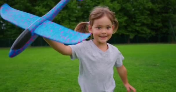 小さな女の子は彼女の手を玩具飛行機で走っています 子供はグリーンフィールドサンセットの周りを走り おもちゃの飛行機を遊んでいます 幼少期の夢はパイロットになる スーパーヒーローの赤ちゃん 子供たちは公園で走っている ハッピーガールパイロット — ストック動画