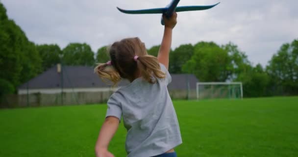 小さな女の子は彼女の手を玩具飛行機で走っています 子供はグリーンフィールドサンセットの周りを走り おもちゃの飛行機を遊んでいます 幼少期の夢はパイロットになる スーパーヒーローの赤ちゃん 子供たちは公園で走っている ハッピーガールパイロット — ストック動画