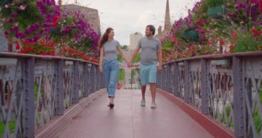 Genç mutlu çift gün batımında köprüde renkli çiçeklerle parkta yürüyor. Modern bir Avrupa şehrinde yürü.