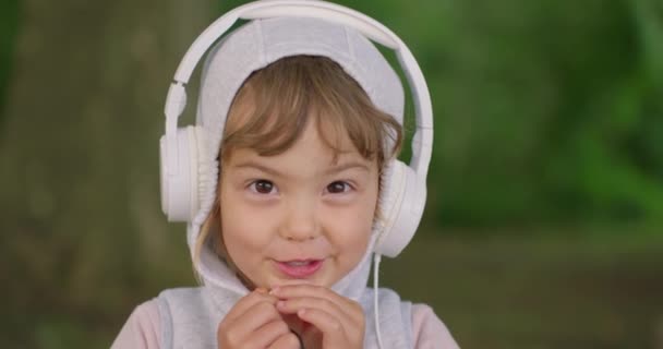 自然の中でヘッドフォンで音楽を聴く3歳の小さなかわいい少女の肖像画 — ストック動画