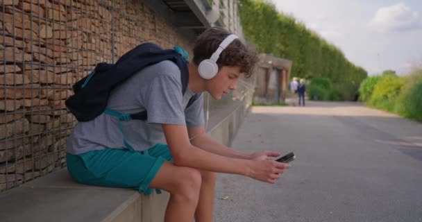 一个可爱的少年坐在公园的长椅上 一边用耳机听音乐 一边看手机 — 图库视频影像