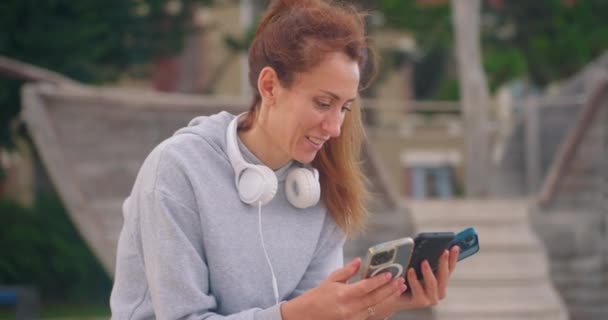 Atrakcyjna Dziewczyna Siedzi Parku Słuchawkach Patrzy Trzy Smartfony Emocje Zaskoczenia — Wideo stockowe
