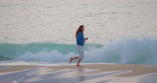 青いスウェットシャツと灰色のパンツのカーリーヘアの少女がサーフラインに沿って走ります 新鮮な空気の中で海の近くでスポーツをする 女性はビーチオーシャンで走っている間 自由で幸せを感じます — ストック動画