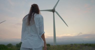 Rüzgar değirmenlerinin arka planında uzun kıvırcık saçlı ve yenilenebilir elektrik üreten bir kadın. Bir mühendis rüzgar enerjisini elektriğe çeviren bir çiftliği inceliyor..