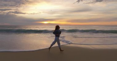 Mavi kazaklı ve gri pantolonlu kıvırcık saçlı bir kız sörf çizgisinde koşuyor. Okyanus kenarında temiz havada spor yapmak. Kadınlar sahilde koşarken kendilerini özgür ve mutlu hissederler..