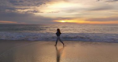Hava görüntüsü. Güzel bir kadın yalınayak koşuyor okyanusta kumsalda akşam vakti inanılmaz bir günbatımı, alacakaranlık.