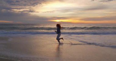 Gün batımında okyanus kıyısında çıplak ayakla koşan bir kadının silueti. Doğayla bütünlük kavramı.