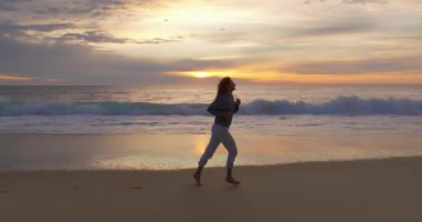 Hava görüntüsü. Güzel bir kadın yalınayak koşuyor okyanusta kumsalda akşam vakti inanılmaz bir günbatımı, alacakaranlık.