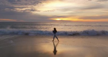 Hava görüntüsü. Güzel bir kadın yalınayak koşuyor okyanusta kumsalda akşam vakti inanılmaz bir günbatımı, alacakaranlık.
