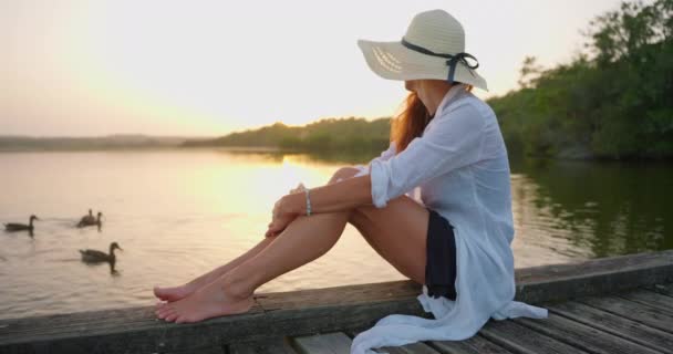 夕日には湖の近くに座っているドレスと帽子の美しい若い女性 休暇を楽しんでいる 座っている女の子 — ストック動画