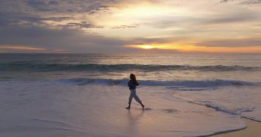 Gün batımında okyanus kıyısında çıplak ayakla koşan bir kadının silueti. Doğayla bütünlük kavramı.