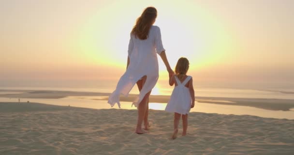 白い服を着たママと娘は 海の日没時に手を握る砂に沿って裸足を歩いている キッズドリームコンセプト 幸せな家族の小さな女の子と自然のリラックスした日没のコンセプト — ストック動画