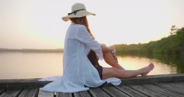 Gün batımında göl kenarında oturan beyaz elbiseli ve şapkalı güzel bir kadın. Tatilin tadını çıkar..