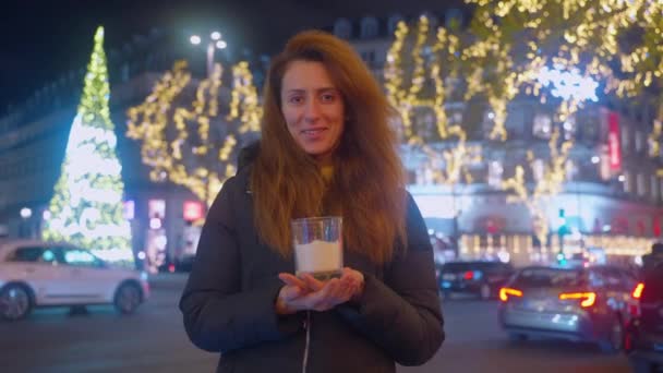キャンドルを持っているカーリーヘアを持つ大人の女性の肖像画 クリスマスの気分 背景にクリスマスの輝く装飾とライトで飾られたパリの通り — ストック動画