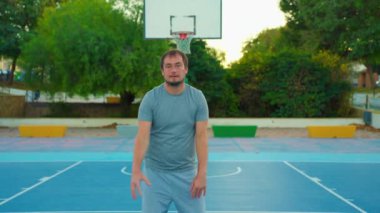 Sporcu dışarıda sahada topla oynuyor. Basketbol potasının arka planında duran ve kameraya bakan adamın portresi. Yavaş çekim sokak basketbolu.