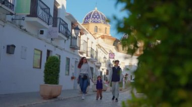 Beyaz evleri olan küçük bir köyde bir İspanyol caddesi boyunca yürüyen bir aile. İspanya 'nın şehir merkezindeki Altea şehrinin mavi çatılı dağ kilisesi. Turistler etrafı geziyor..