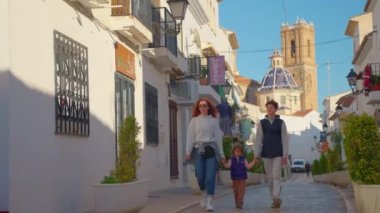 Beyaz evleri olan küçük bir köyde bir İspanyol caddesi boyunca yürüyen bir aile. İspanya 'nın şehir merkezindeki Altea şehrinin mavi çatılı dağ kilisesi. Turistler etrafı geziyor..