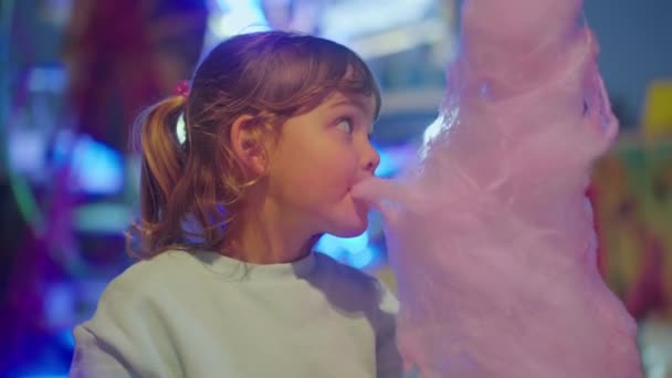 小さなかわいい女の子は 遊園地の背景にコットンキャンディーを食べます 子供の頃のコンセプト ケアフリーと楽しい — ストック動画