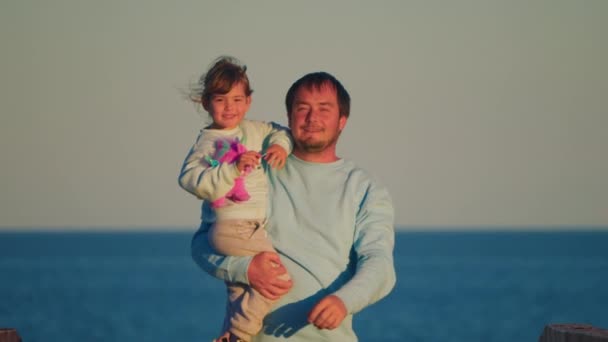 父親は日没時に青い海の背景に彼の腕にかわいい娘を運んでいる 子供や家族と過ごした幸せな父親の概念 — ストック動画
