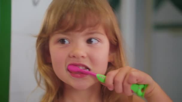 小さなかわいい少女は 鏡で自分自身を見ている歯ブラシで彼女の歯を希釈的に磨きます クローズアップ — ストック動画