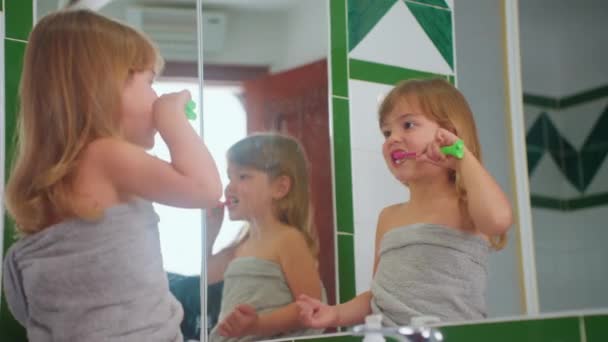 귀여운 소녀는 부지런히 거울에서 자신을보고 칫솔로 그녀의 이빨을 닦습니다 — 비디오