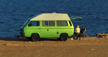 Köpekli bir çift, denizin üzerindeki bir uçurumda eski yeşil karavanın yanında dinleniyor. Aile tatili karavanı, karavan tatili, karavan tatili..