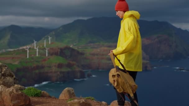 穿着黄色夹克和背包的迷人的女游客看着田野里的风车 — 图库视频影像