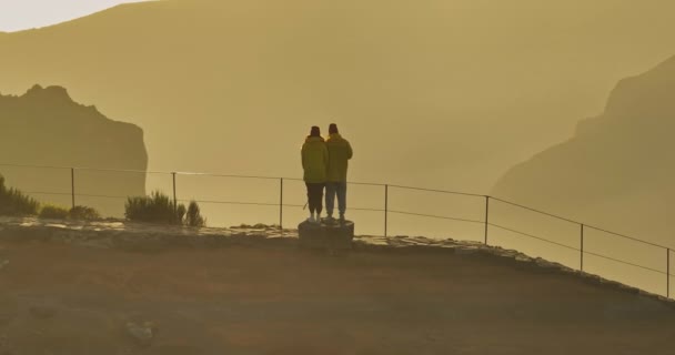 バックパックのスローモーションウォーキングと2人の観光客のチームワークは 日没の太陽の光の輝きでシルエットを行きます カメラは旅行や冒険のカップルをフォローしています 美しい山の谷で家族の散歩 — ストック動画