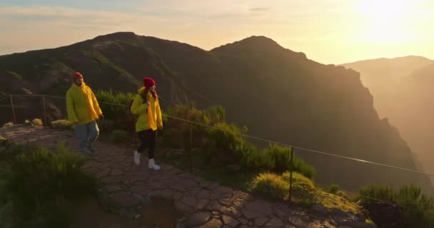 バックパックのスローモーションウォーキングと2人の観光客のチームワークは 日没の太陽の光の輝きでシルエットを行きます カメラは旅行や冒険のカップルをフォローしています 美しい山の谷で家族の散歩 — ストック動画