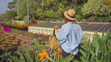 Gezgin, Madeira ve Funchal şehrinin çeşitli bitki örtüleriyle birlikte tarihi bir bahçeyi ziyaret ediyor. Botanik bahçesinde renkli çiçekler arasında yürüyen şapkalı ve sırt çantalı bir kadın..