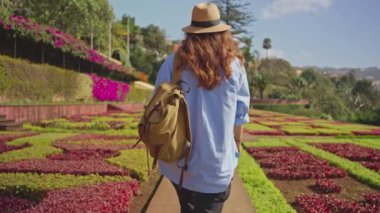 Gezgin, Madeira ve Funchal şehrinin çeşitli bitki örtüleriyle birlikte tarihi bir bahçeyi ziyaret ediyor. Botanik bahçesinde renkli çiçekler arasında yürüyen şapkalı ve sırt çantalı bir kadın..