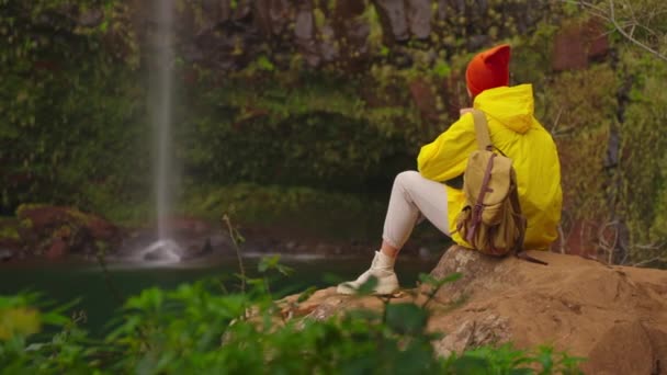 高地の滝近くの黄色いレインコートで女性の旅行者は自然と人生を楽しんでいます 山を旅する 旅行の冒険 ライフスタイルコンセプト イエローパッファージャケットのトラベラー — ストック動画