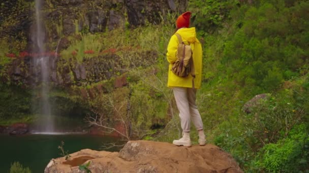 高地の滝近くの黄色いレインコートで女性の旅行者は自然と人生を楽しんでいます 山を旅する 旅行の冒険 ライフスタイルコンセプト イエローパッファージャケットのトラベラー — ストック動画