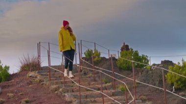 Turistik kıyafetli, sarı ceketli ve sırt çantalı bir kadın destansı dağ zirvesinde yürüyor..