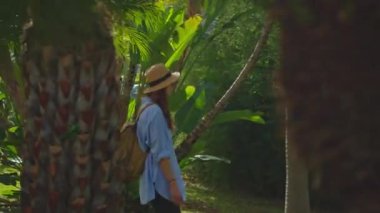 Kamera, şapkalı ve sırt çantalı gezgin kadını takip ediyor. Ormandaki tropikal bahçede ağır çekim kadın yürüyüşü..