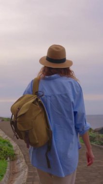 Dikey video. Mutlu genç bayan turist okyanus boyunca şapka ve sırt çantasıyla yürüyüp en güzel gün batımında okyanus manzarasının keyfini çıkarıyor. Madeira Adası, Portekiz.