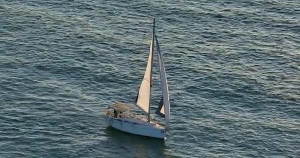太陽の光線に黒い帆で白いヨットの空中撮影を確立する 水の上に輝いている ドローンから撮影された青い海のセーリングヨットのシルエット — ストック動画