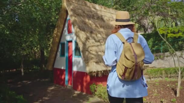 女の子は帽子とバックパックで旅行し ポルトガルマデイラのサンタナの特別な伝統的なA字型の家を訪問します — ストック動画