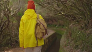 Sarı ceketli bir kız sırt çantasıyla seyahat eder ve Madeira, Levada dos Tornos, Portekiz 'deki sıradışı yerleri ve yolları ziyaret eder..