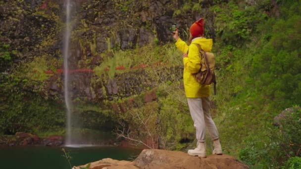 노란색 재킷과 배낭에있는 여행자는 아름답고 수없는 폭포에 도달하여 전화에 사진을 — 비디오