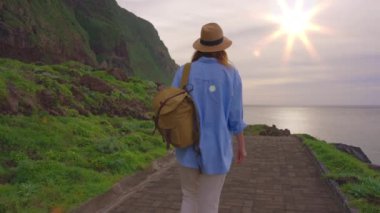 Mutlu genç kadın turist, gözlem güvertesinde şapka ve sırt çantasıyla okyanus boyunca yürüyor ve en güzel günbatımında okyanus manzarasının keyfini çıkarıyor. Madeira Adası, Portekiz.
