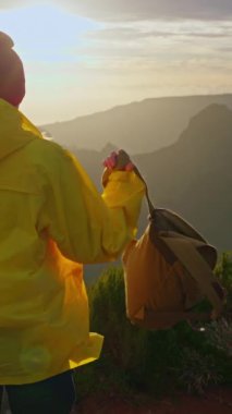 Dikey video. Sarı ceketli ve sırt çantalı kadın gün batımında dağların inanılmaz manzarasına bakıyor. Yakın çekim..