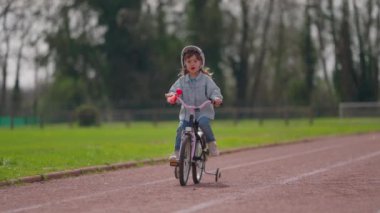 Koruyucu kasklı küçük tatlı kız stadyumda yedek tekerlekleriyle bisiklet sürüyor. Güneşin ve bahar havasının tadını çıkarıyor..
