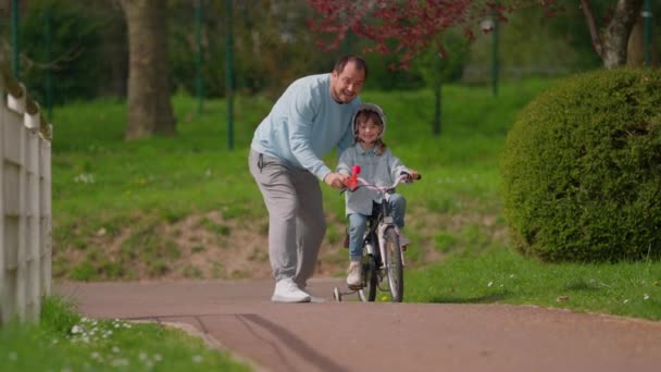 小女孩和细心的父亲在公园里骑车 父亲正在教她骑自行车 当她成功的时候 她很高兴 幸福的年轻家庭 父亲和童年 积极的生活方式概念 — 图库视频影像